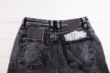 Beyaz arka plana karşı gri kot pantolon cebinde dolar ve akıllı telefon. Yakın plan..