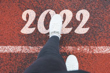 Mutlu yıllar 2022. Kadınların bacakları beyaz çizgiyi geçer ve 2022 'ye adım atar. Koşu bandındaki beyaz spor ayakkabıların 2022 nolu metni, yeni yıl kutlaması konsepti.
