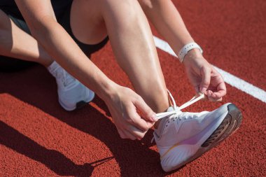 Spor ayakkabı bağcıklarını eğitirken bağlayan kadın. Spor kıyafetli bir kadın ayakkabı bağı bağlayıp çömeliyor. Açık hava eğitim stadyumunda gri gardırop ve beyaz spor ayakkabılar, kadın vücudunun yakın çekimi.