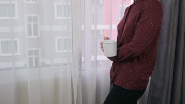 Молодая женщина стоит у окна и пьет кофе.. — стоковое видео