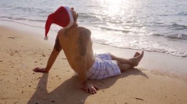 Denizin kenarında Noel Baba şapkası takan bir adam oturuyor. - Mutlu yıllar. Yüksek kalite 4k görüntü