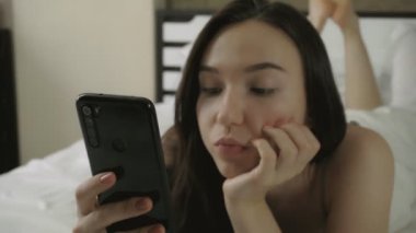 Genç ve güzel bir kadın yatağa uzanıyor ve akıllı telefon kullanıyor.