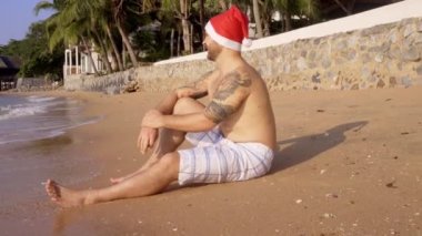 Denizin kenarında Noel Baba şapkası takan bir adam oturuyor. - Mutlu yıllar. Yüksek kalite 4k görüntü