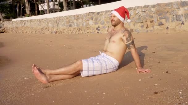 Homem Senta Areia Junto Mar Com Chapéu Pai Natal Feliz — Vídeo de Stock