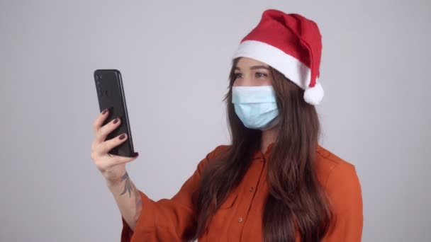 Tıbbi maskeli bir Noel Baba şapkası takan ve telefonuyla görüntülü konuşma yapan bir kadın. — Stok video