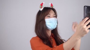 Saçında Noel süsleri olan ve tıbbi maske takan bir kadın bir telefon tutuyor ve video aracılığıyla iletişim kuruyor. Koronavirüs salgınının ortasında kutlama. Yüksek kalite 4k görüntü