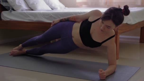 Jovem deitada em um tapete esportivo no quarto e fazendo exercícios de prancha lateral — Vídeo de Stock