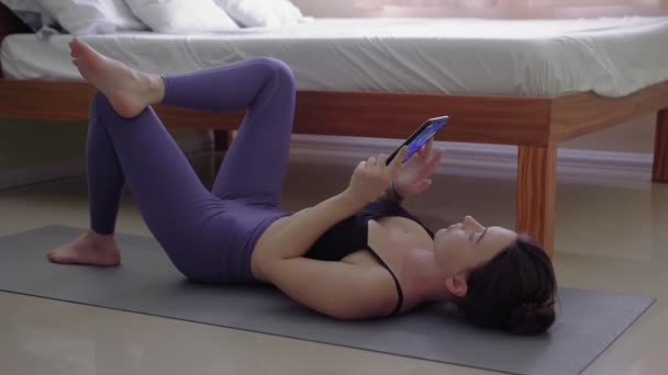 Молодая женщина лежит на спортивном ковре в спальне и сёрфит телефон — стоковое видео
