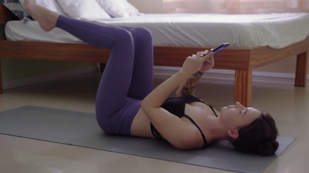 若い女性がベッドルームでスポーツラグに横たわっていると電話でサーフィン — ストック動画