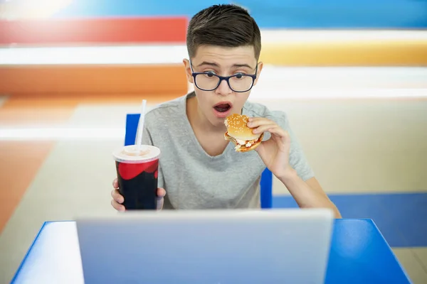 Caucasian white teenager eating burger and looking surprised at laptop — Stock Photo, Image