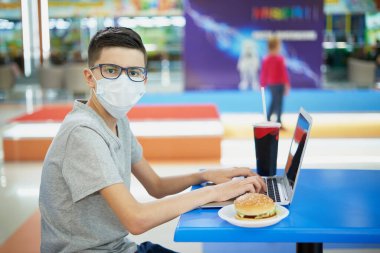 Yemekte yemek katında dizüstü bilgisayarı olan maskeli bir çocuk kameraya bakar.