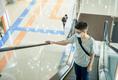 Alışveriş merkezinde yürüyen merdivende maske takan genç bir çocuk.