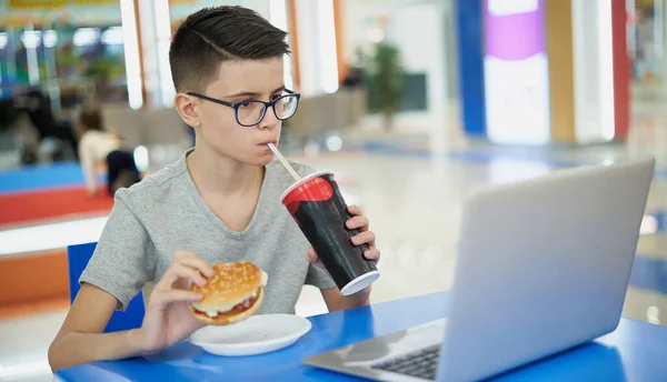 Хлопчик-підліток їсть нездорову фаст-фуду і п'є соду, сидячи в продовольчому дворі — стокове фото