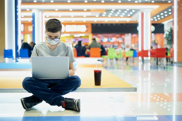 Magányos tinédzser fiú arcmaszkban laptopot használ a plázában — Stock Fotó