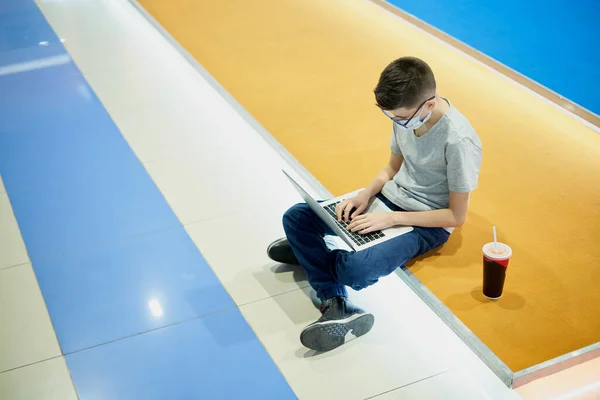 Adolescente menino na máscara facial sentado no chão e usando laptop — Fotografia de Stock