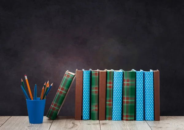 pile of books in colorful covers and pencils in holder on wooden table with green blackboard background. Distance home education. Back to school, quarantine concept of stay home
