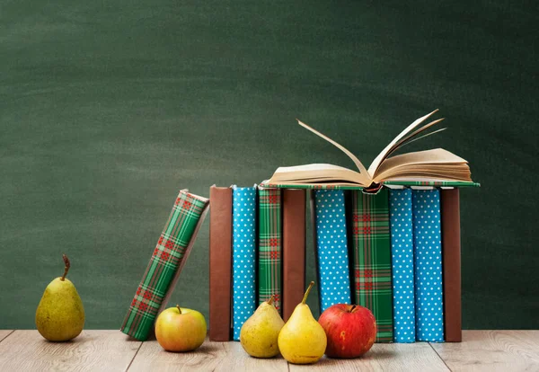 Açık ders kitabı, renkli kapaklar içinde kitap yığını, ahşap masa üzerinde yeşil karatahta arka planlı armut ve elmalar. Uzaktan eğitim. Okula dönmek, karantina konsepti evde kalmak.