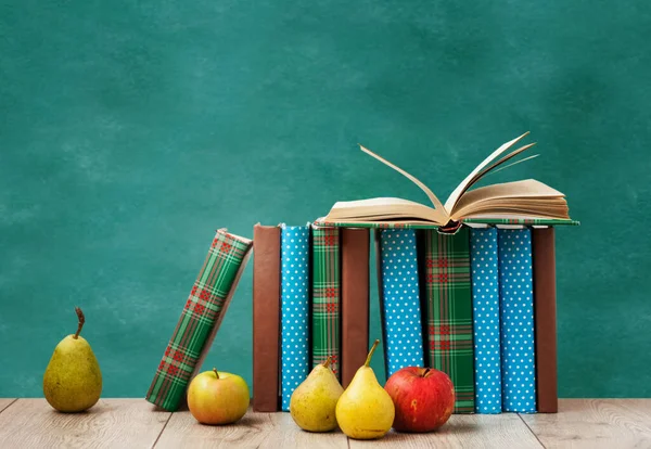 Open textbook, pile of books in colorful covers, pears and apples on wooden table with green blackboard background. Distance home education. Back to school, quarantine concept of stay home