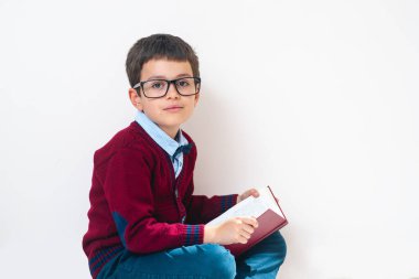 Süveter ve gözlüklü okul çocuğu elinde açık bir kitapla oturuyor ve bize bakıyor. Kavramsal. Boşluğu kopyala.