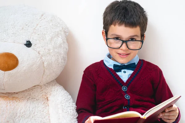 The schoolboy in a sweater and glasses sits next to a large teddy bear with an open book in his hands and looks at us. Conceptual. Copy space.