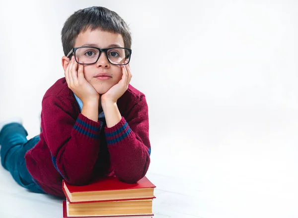 The schoolboy in a sweater and glasses lies with his hands on a stack of books and looks at us. Conceptual. Copy space.