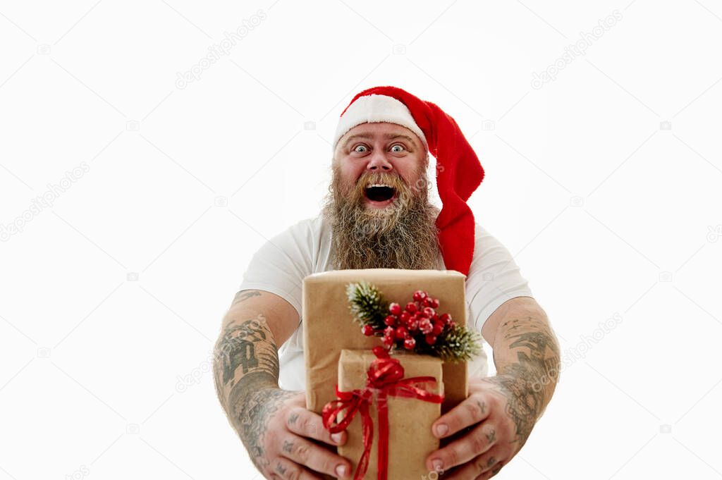 Isolated portrait on a white background of an overweigh laughing man with tattooed arms in Santa hat presenting Christmas gifts at camera