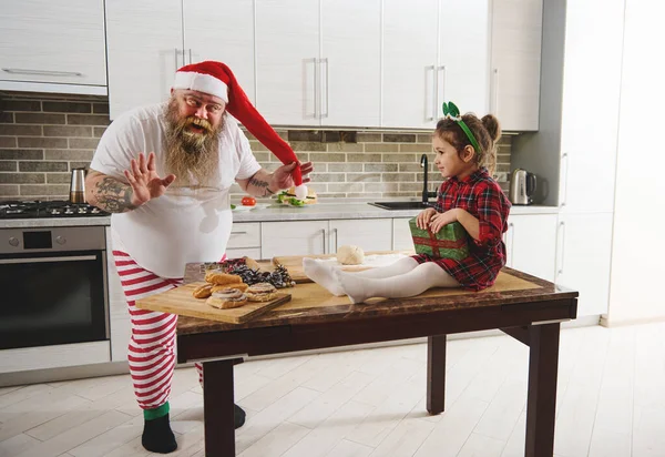 A cheerful father in a Santa hat entertains his young daughter sitting on a table and holding a gift in her hands. Having fun holidays