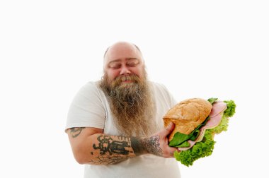 Kolları dövmeli, mutlu, obez sakallı bir adam kocaman bir burger tutuyor ve gözleri kapalı gülümsüyor..