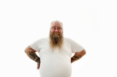 Kolları dövmeli, kilolu sakallı bir adam ellerini kemerinde tutuyor.