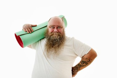 Kolları dövmeli, omuzunda spor paspası tutan ve kameraya gülümseyen kilolu sakallı bir adam..