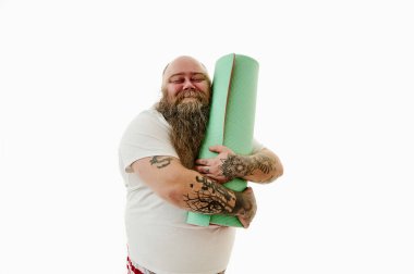 Kolları dövmeli mutlu şişman bir adam spor paspasına nazikçe sarılıyor ve gözleri kapalı gülümsüyor. Beyaz arkaplanda izole edilmiş bir portre