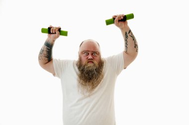 Aşırı kilolu sakallı, kollarında dambıllarla egzersiz yapan ve kameraya ciddi ciddi bakan bir adam.