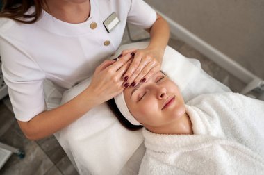 Güzeller güzeli genç bir kadın masaj masasında dinlenirken güzellik uzmanı SPA salonunda yüz masajı yapıyor.