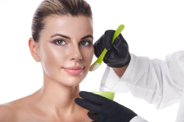 Güzellik uzmanının elleri yeşil spaluta ve beyaz güzellik ürünleriyle dolu bir kase tutuyor ya da beyaz tenli güzel bir kadının yüzünün yanında krem sürüyor. Beyaz arkaplanda izole edilmiş portre