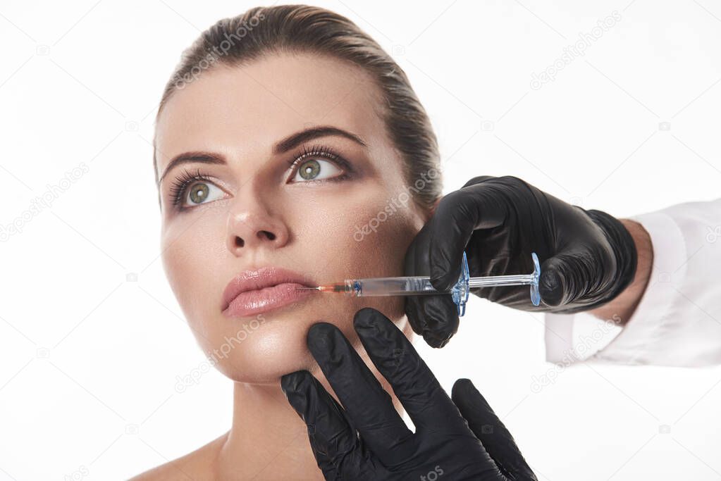 Lips Augmentation Concept. Hands in black gloves making injection of filler to attractive woman's lips. Portrait on a white background.