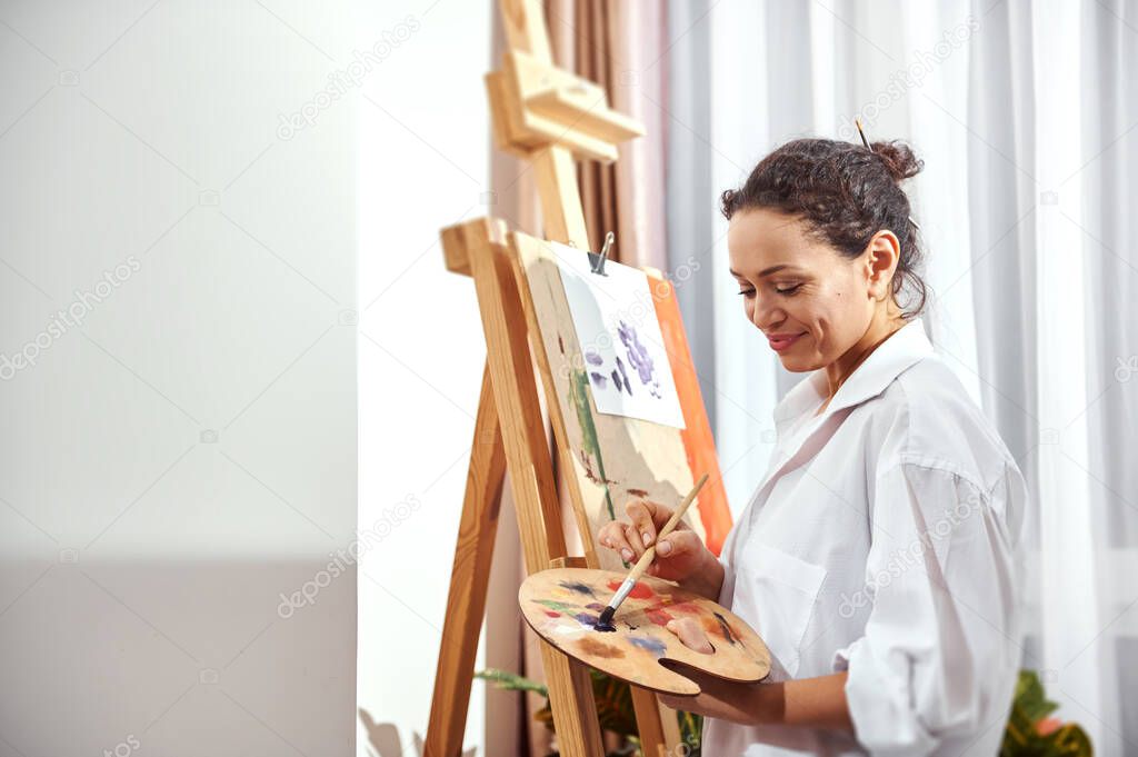 Side view of a smiling happy female painter mixing oil paint on a palette and standing in front of an easel