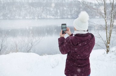 Kalın elbiseli bir kadının arkasından bir akıllı telefon tutarken ve kar yağarken güzel bir kar manzarası çekerken görüntüsü..