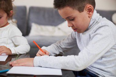 Renkli kalemle çizmeye odaklanmış gri gömlekli bir çocuğun yan görüntüsü. Masada duran renkli tahta kalemler..