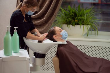 Siyah üniformalı bir kuaför ve koruyucu bir tıbbi maskeyle müşterinin saçını güzellik salonunda yıkarken. Salgın sırasında güzellik salonlarının yeniden açılması kavramı