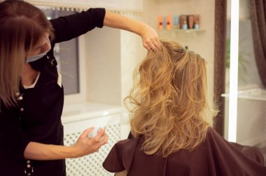 Koruyucu tıbbi maskeli kadın kuaförü ve siyah üniformalı saç spreyi kullanan ve güzellik salonundaki sarışın kıza saç stili yapan.