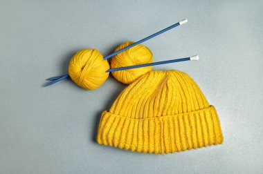 Sarı örülmüş yün bir şapka ve gri arkaplandaki uzun mavi örgü iğnelerine sarılı iki yün iplik.