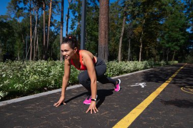 Koşucu kadın, açık hava koşusunda başlangıç pozisyonunda. Arka planda güzel bir park.