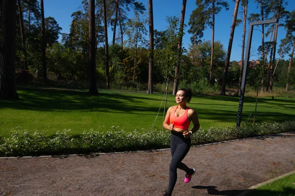 The young woman in sportswear runs in the park against a background of green grass. Outdoor sports.