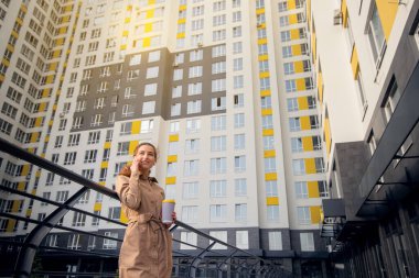 Genç, mutlu ve başarılı bir kadın elinde bir fincan kahveyle yüksek binanın arka planında telefonla konuşuyor.