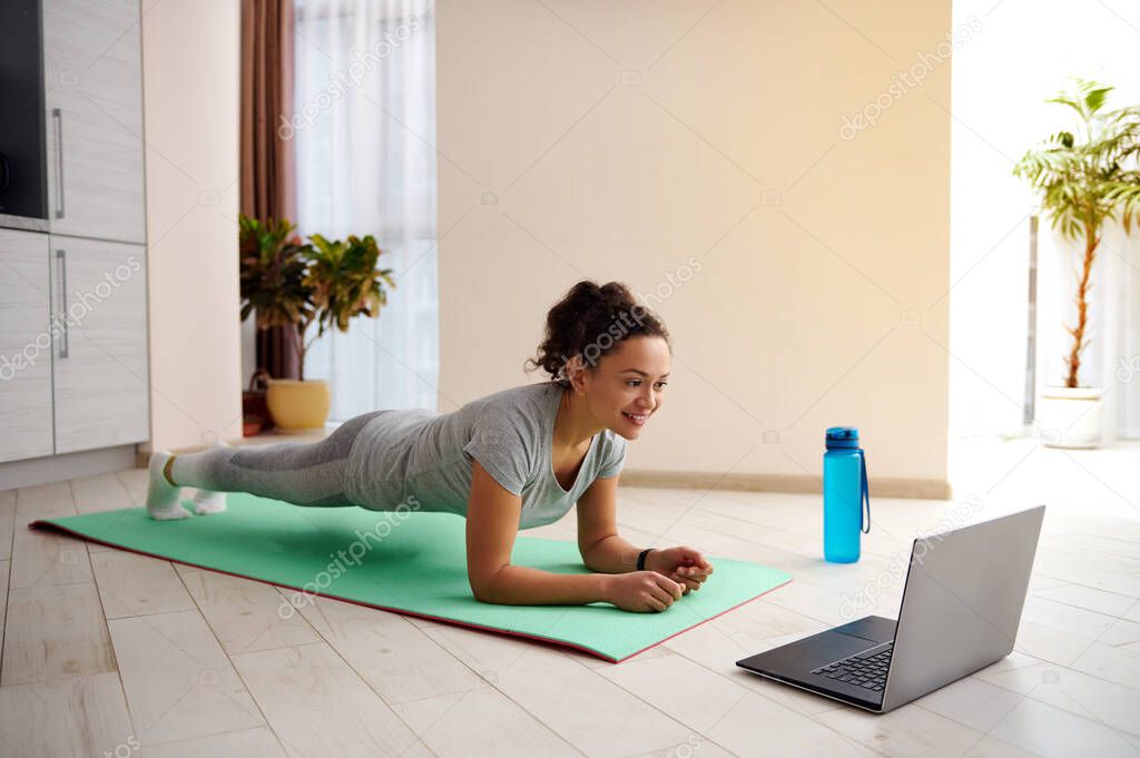 Cute woman fitness instructor performing plank exercise on a yoga mat via laptop. Stay home and quarantine workout concept