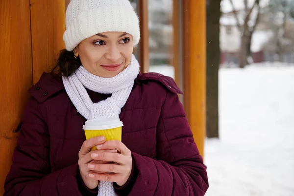 Young woman wearing white scarf and white woolen hat stands with a coffee to go at a winter snowy park.
