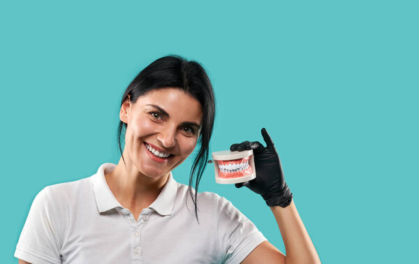 Красивая молодая доктор держит челюсть или модель человеческих зубов рядом с лицом и улыбается, глядя в камеру. Портрет изолирован на синем фоне