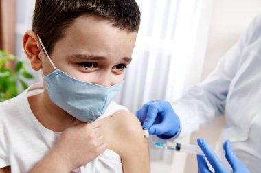 Tıbbi maske takan korkmuş bir çocuğun portresi, ona aşı yapan doktorun elleri.