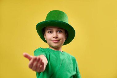 Leprikon yeşil İrlanda şapkalı küçük çocuk avucunu kameraya uzatıyor. Aziz Patrick Günü konsepti. Boşluğu ve sarı arkaplanı kopyala