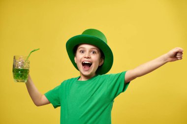 Yeşil tişörtlü ve cüce şapkalı mutlu çocuk elinde yeşil içkili bir bardak tutuyor ve Aziz Patrick Günü 'nü kutlayarak mutluluğunu ifade ediyor.